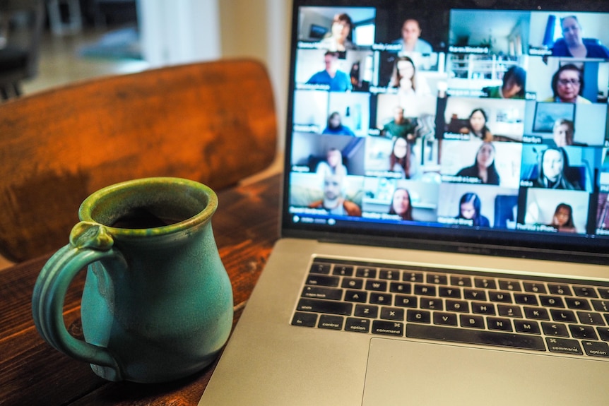Laptop which has more than a dozen people on the screen in a zoom meeting. A bright teal mug with a yellow rip sits to the left.