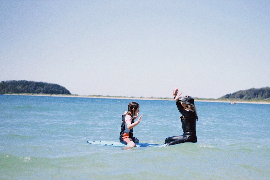 Una niña y una mujer están en el agua sobre una tabla de surf, una frente a la otra con los brazos en alto. 