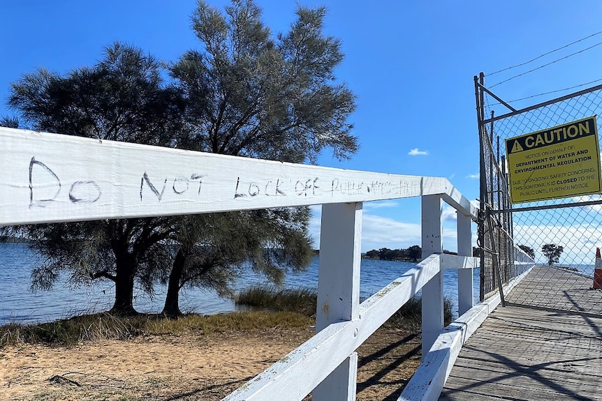 A close up shot of a section of a timber jetty, with white railing. Some graffiti is evident, as is an area that is fenced off.