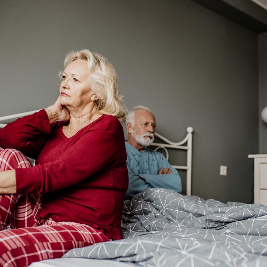older woman looking unhappy wearing red sits on a bed with a man in the background crossing his arms