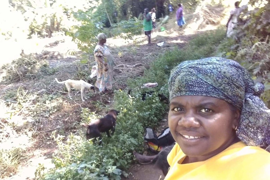 A woman takes a selfie while standing next to a field with other people standing and sitting nearby.