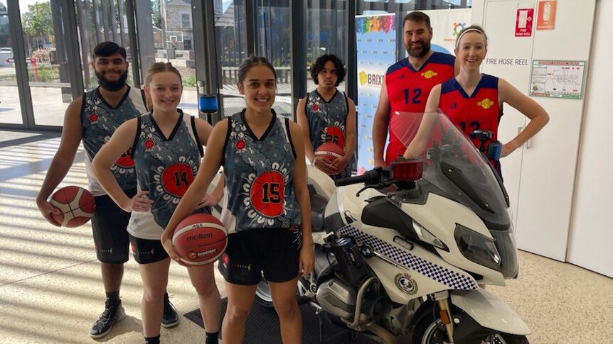 aboriginal boys and girls in basketball uniforms with balls pose near police bike with two adults in red basketball uniforms
