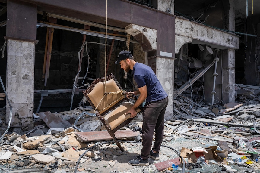 An Arab man in a cap carries a chair through rubble 