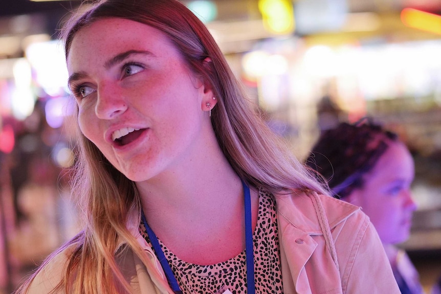 Keely Johnson looks to the left and smiles as she stands inside a bowling alley. She wears a tan jacket and leopard print top.