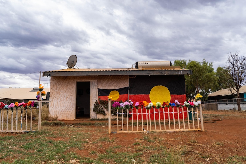 A small house with an Aboriginal flag painted on the exterior and flowers lining the fence. 
