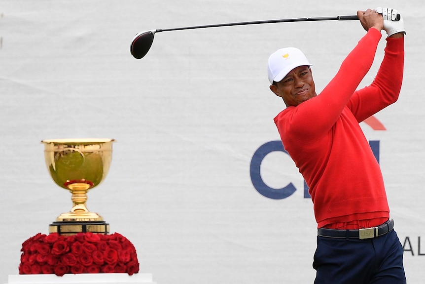 Tiger Woods plays a shot wearing a red jumper in front of the golden President's Cup