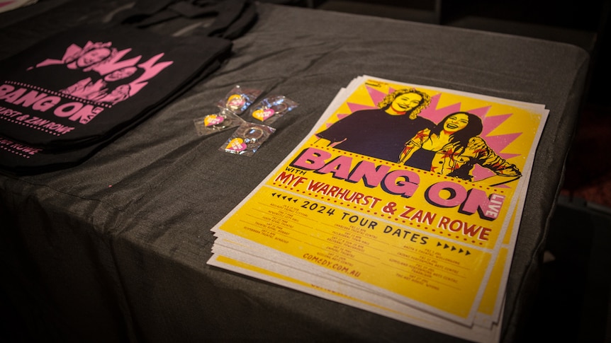 A stack of yellow posters saying 'Bang On Live, Myf Warhurst and Zan Rowe' on a merch desk
