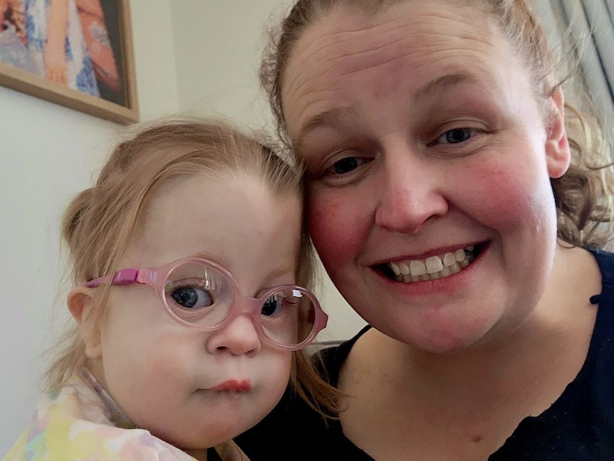 A mother holds a toddler wearing glasses in her arms and smiles.