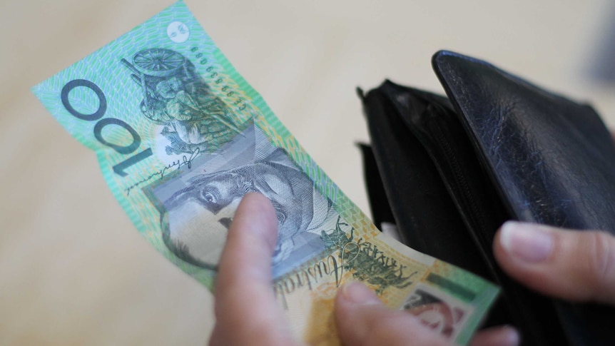 A hand holding a $100 note next to a wallet