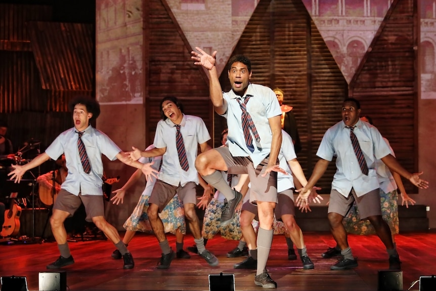 Group of young Indigenous men in school uniforms dancing on brightly lit stage in formation.