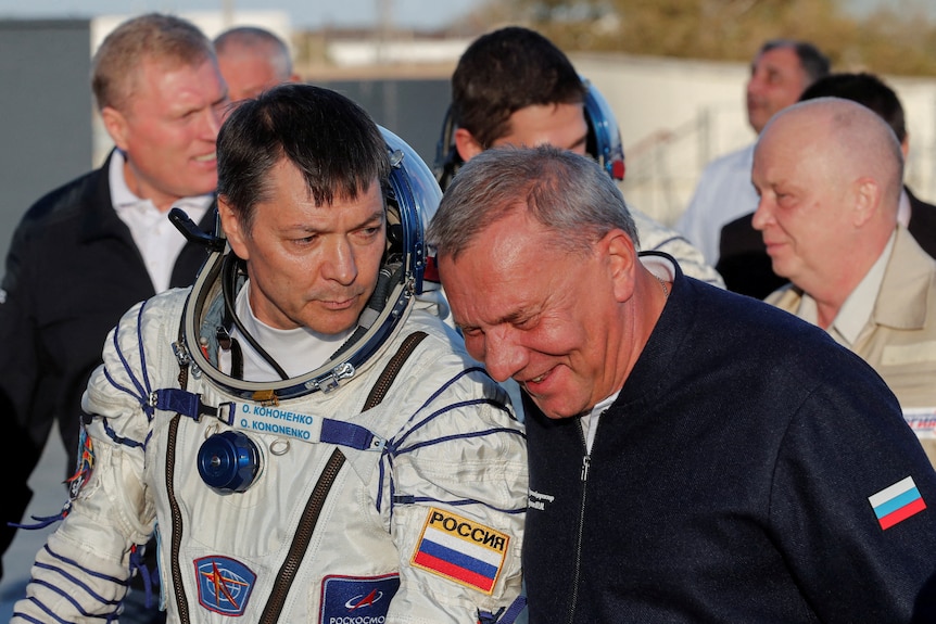 A middle-aged man in an astronaut suit stands next to an older man in a tracksuit bearing the Russian flag.