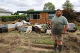 A man standing in front of his brick, flood damaged home with all his possessions strewn over the lawn. 