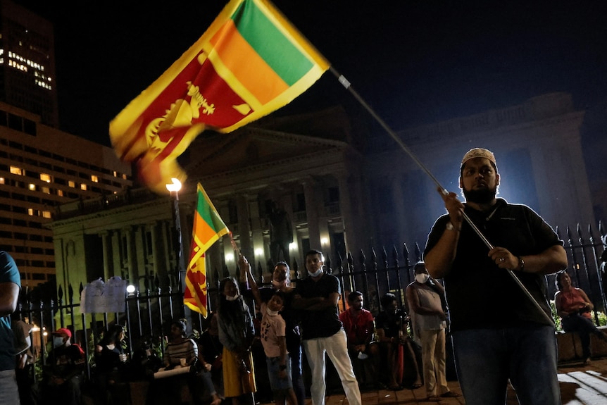 A man is backlit by a spotlight as he waves the Sri Lankan flag amid a crowd at night time
