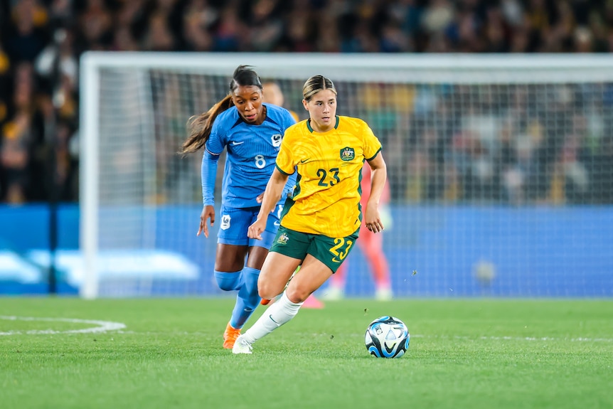 A soccer player wearing yellow and green kicks the ball during a game with a player wearing blue behind her