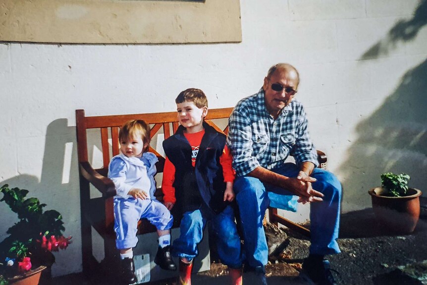 An older man wearing sunglasses and a flannel shirt sits on wooden bench next to two small children, smiling.