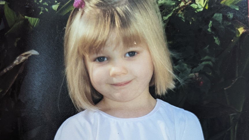 A portrait of blonde toddler with a pink clip in her hair, white top, smiling slightly.