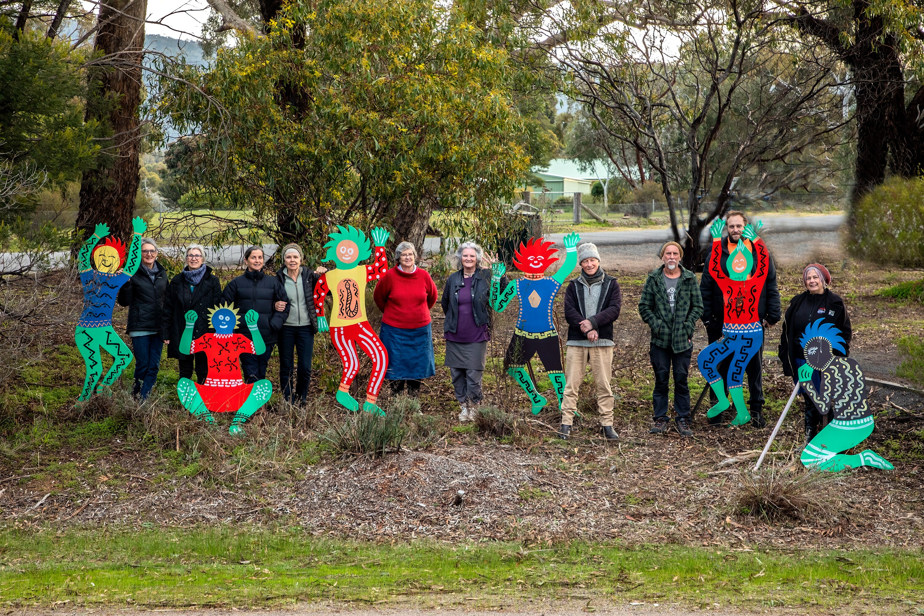 A group of people stand on mulch between sculptures of colorful people with trees in the background. 