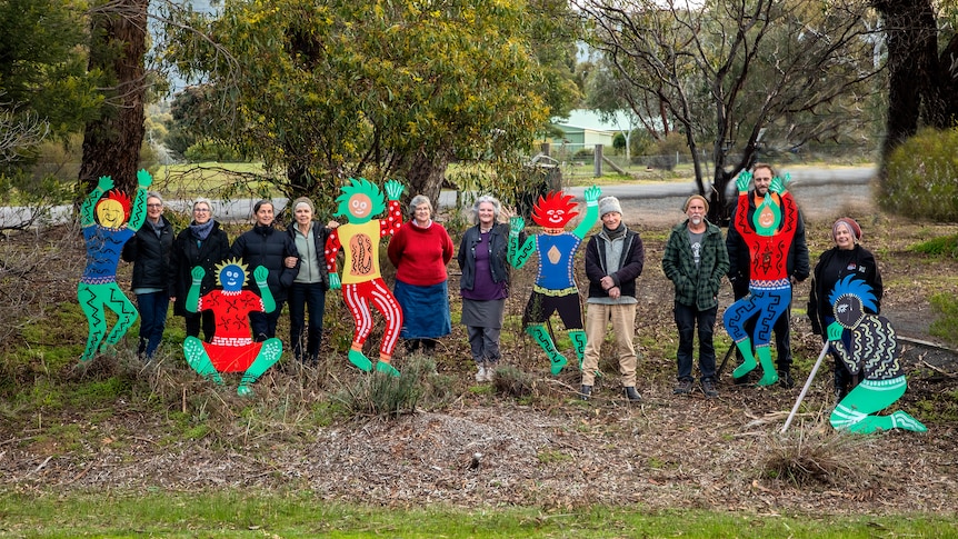 A group of people stand on mulch between sculptures of colourful people with trees in the background. 