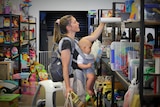 A woman with a baby strapped to her chest peruses a shelf of toys. 