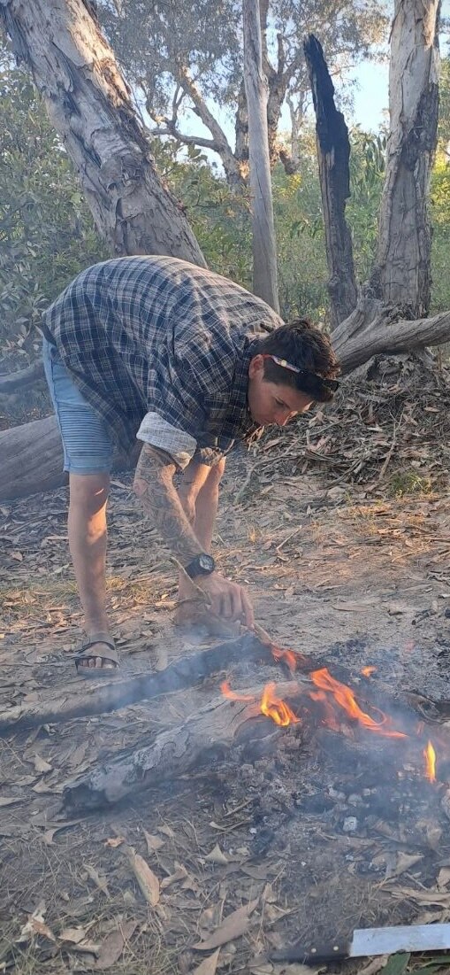 A woman wearing a checked flannelette leans over a campfire and tends to it.