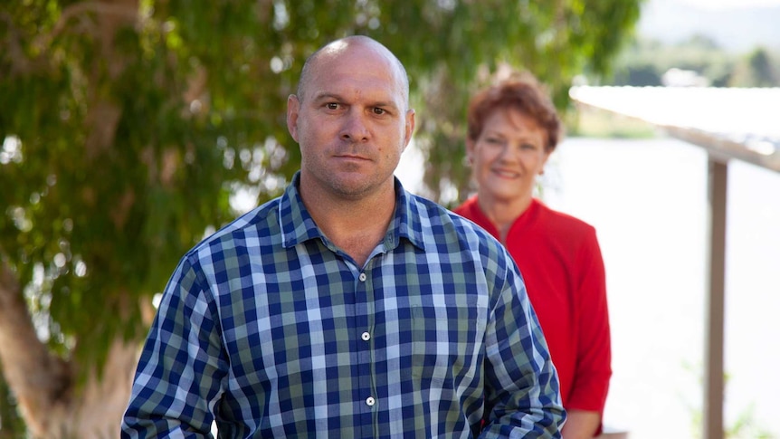 An unsmiling bald man in a blue checked shirt stands in front of a tree, with a woman behind him