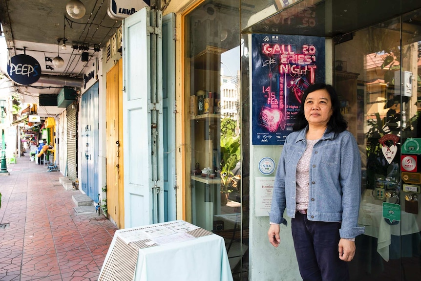 A woman with black hair wearing a denim jacket and black pants stands in front of a restaurant on a street.