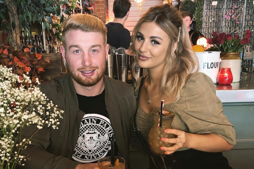 A man at a restaurant table, holding a drink,  sat next to a blonde-haired woman also a drink.