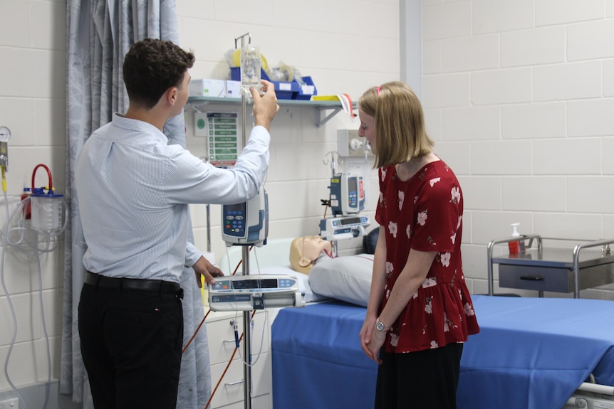 A man with his back to the camera handles an IV drip with a woman looking on, hospital bed behind them