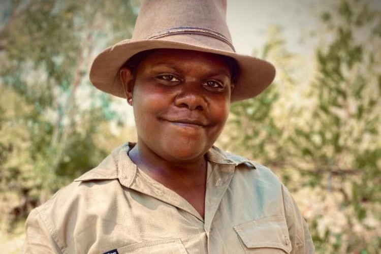 A smiling Aboriginal woman in khaki and a wide-brimmed, bush behind her.