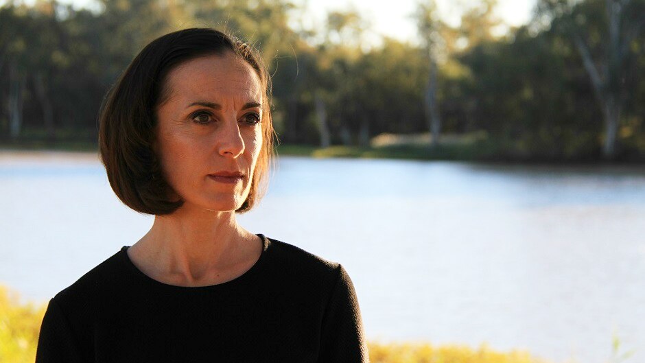 A woman stares into the distance, against a backdrop of a river with lush green banks.