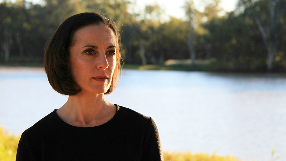 A woman stares into the distance, against a backdrop of a river with lush green banks.