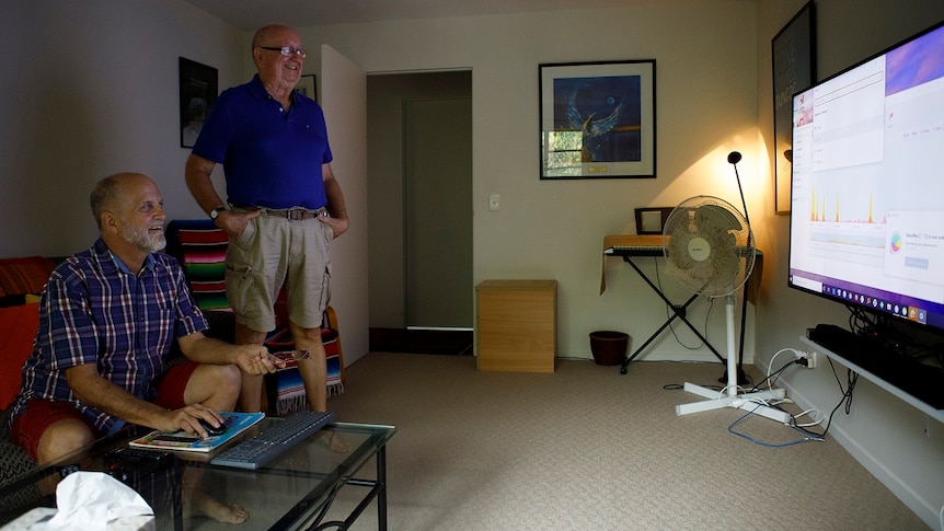 Two men stand looking at a large TV screen in a room of their house for a story on share housing when retired.