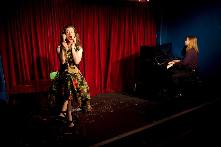 Bethany on stage in character while a man plays a piano.