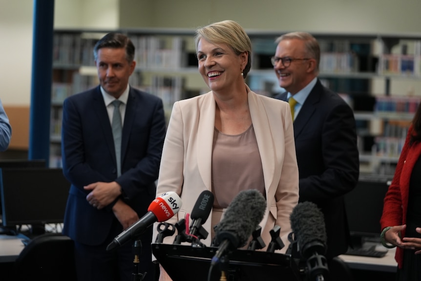 A woman in a neutral business suit smiles in front of a bank of microphones