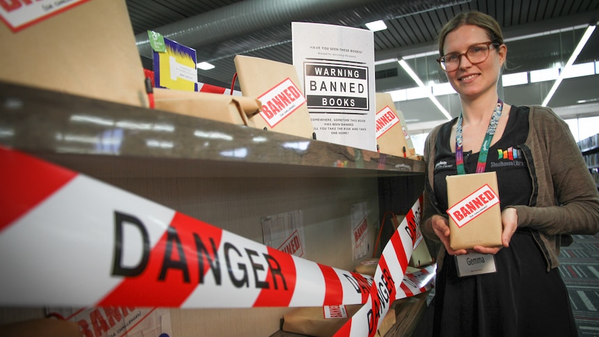 A woman stands in the library holding a book wrapped in brown paper