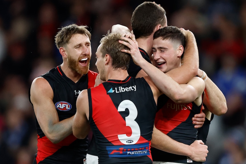 A group of smiling Essendon AFL players gather together and hug each other after a win. 