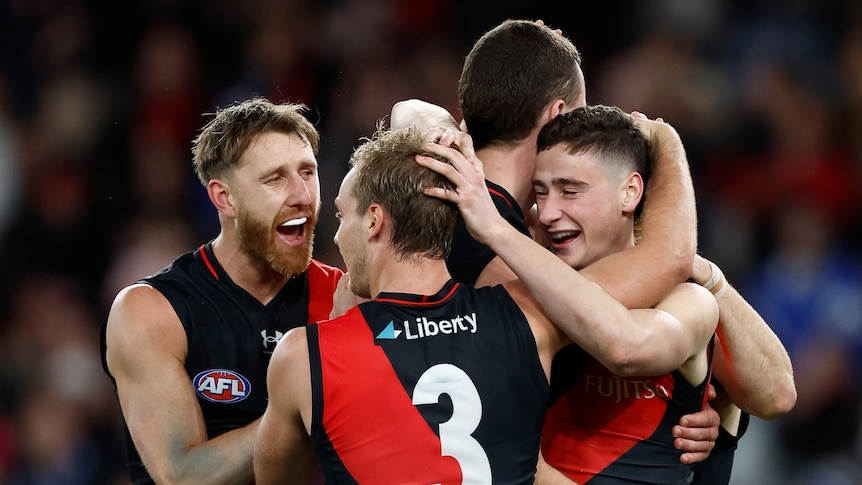 A group of smiling Essendon AFL players gather together and hug each other after a win. 