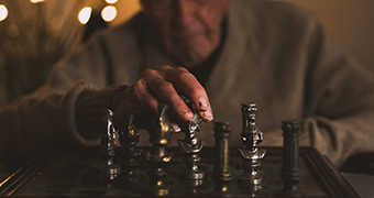 A very old man moves a chess piece on a board in low light.