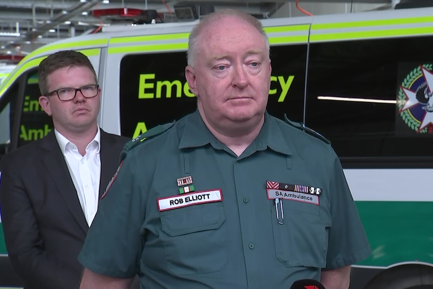 A man in a green paramedic uniform standing in front of another man and an ambulance