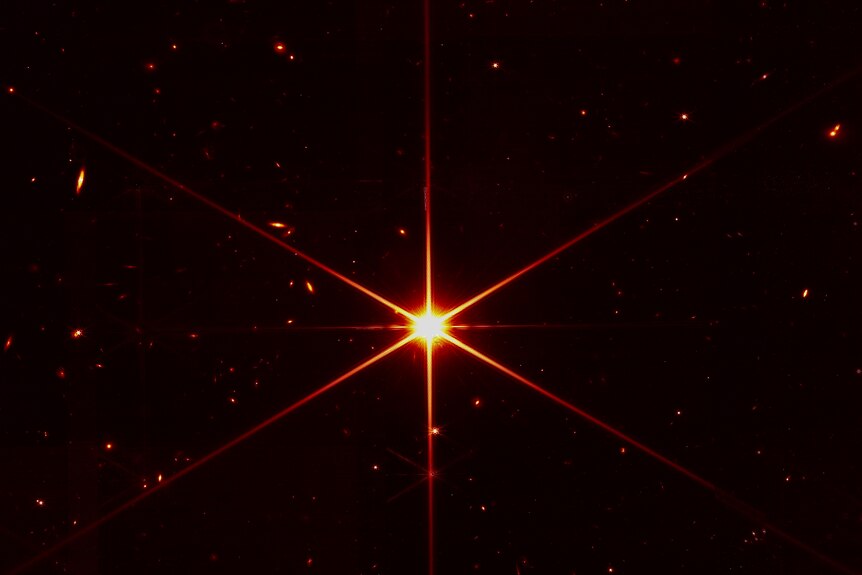 A red star with six lines coming out from the center with other galaxies and stars surrounding it.