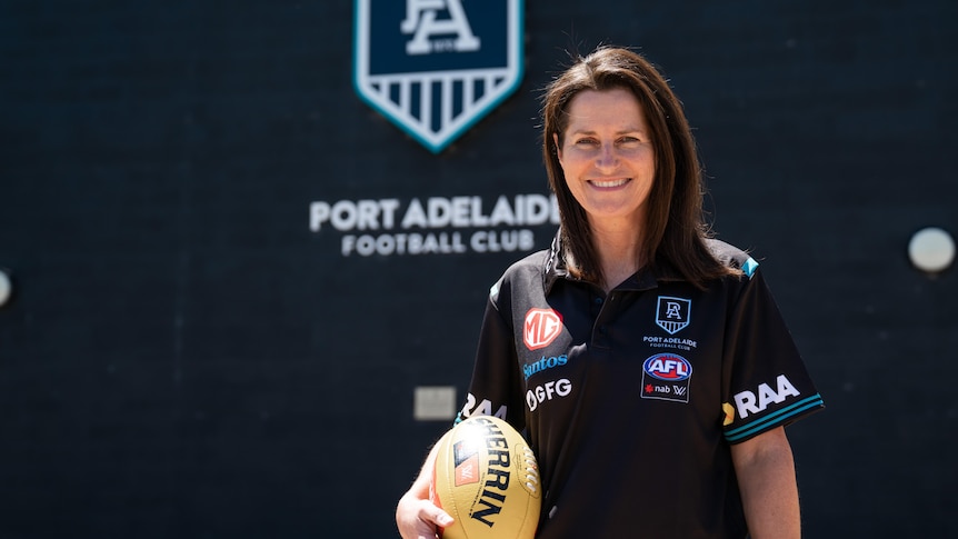 A woman in Port Adelaide Football Club polo shirt holding a football with club logo in the background