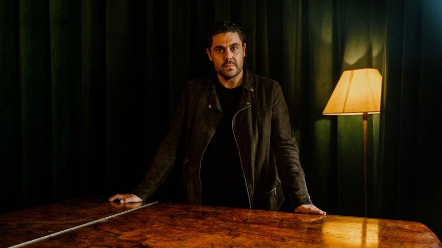 Dan Sultan stands, arms wide on a glazed wooden table with a lamp and green curtain behind him