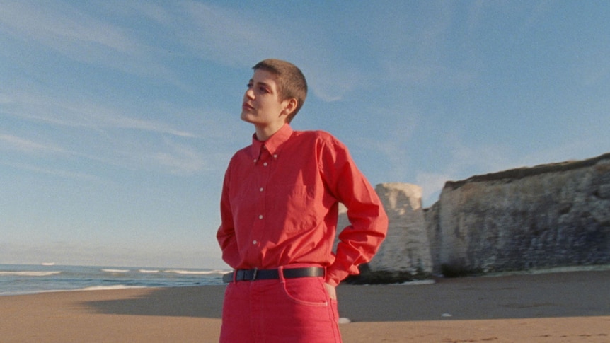 Porridge Radio singer Dana Margolin stands on a beach looking into the distance, wearing a pink suit
