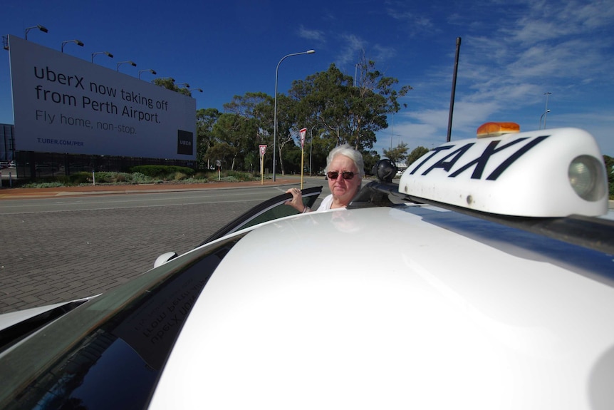 Pat Hart stands at the driver's side door of her car with a taxi sign on its roof and an Uber road sign in the background.