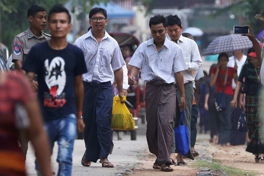 Two men walk among a small crowd wearing traditional Myanmarese wrapped skirts with dress shirts on top, carrying plastic bags.