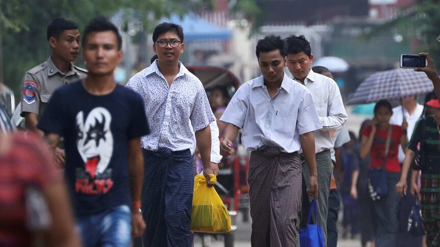 Two men walk among a small crowd wearing traditional Myanmarese wrapped skirts with dress shirts on top, carrying plastic bags.