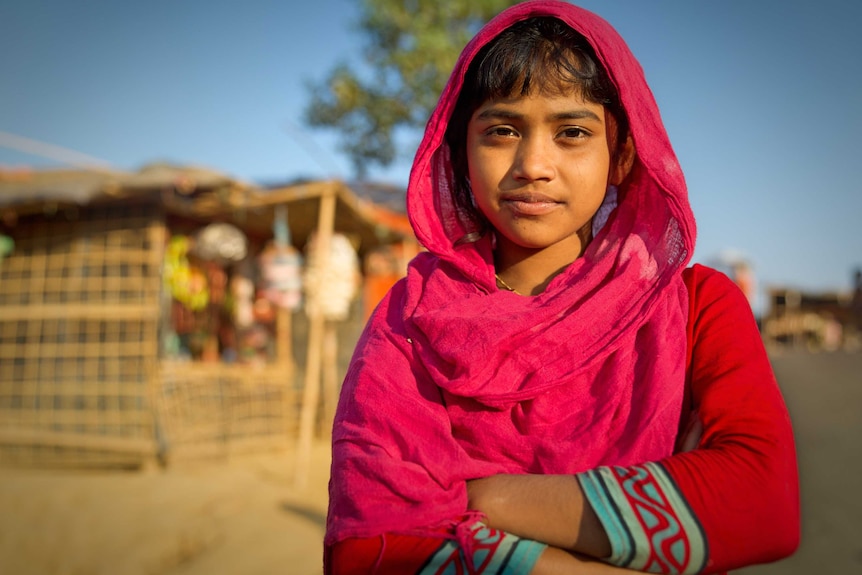 A Rohingya girl in pink clothes poses for a photo with her arms crossed.