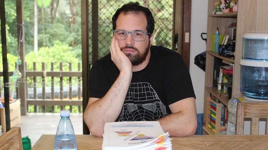 Man with dark hair and beard, wearing glasses holding face in his hands sitting at a desk in front of pile of papers.