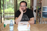 Man with dark hair and beard, wearing glasses holding face in his hands sitting at a desk in front of pile of papers.