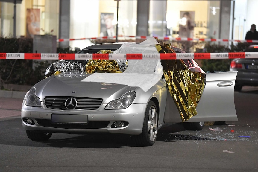 A silver car shows signs of damage, with windows shot out and door open. It is surrounded by police tape.